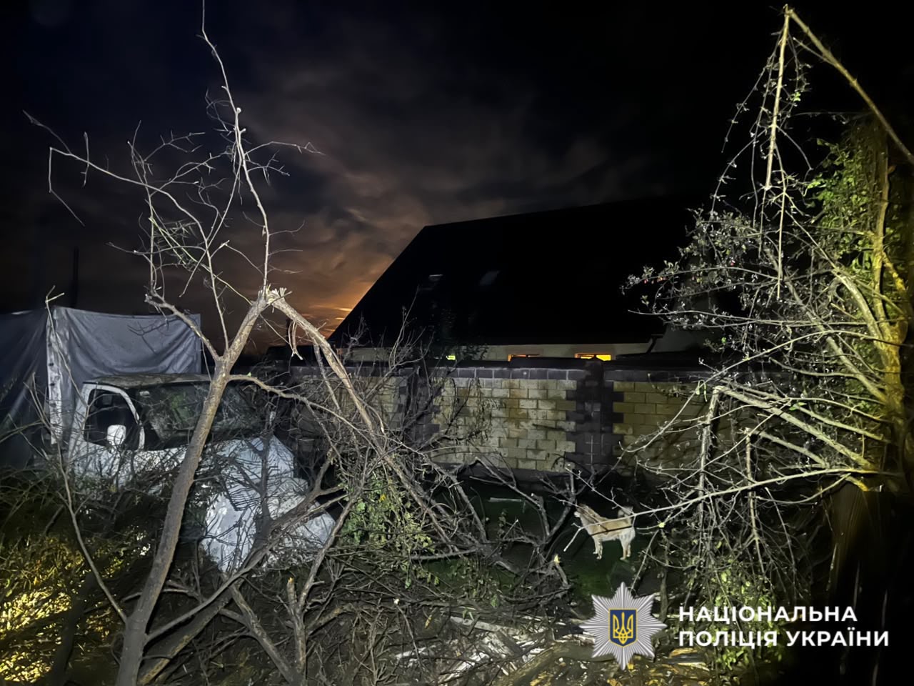 Ніч, освітлена порушена будівля з пробитим вікном та пошкодженою дахом, оточена голими гілками дерев після удару дроном