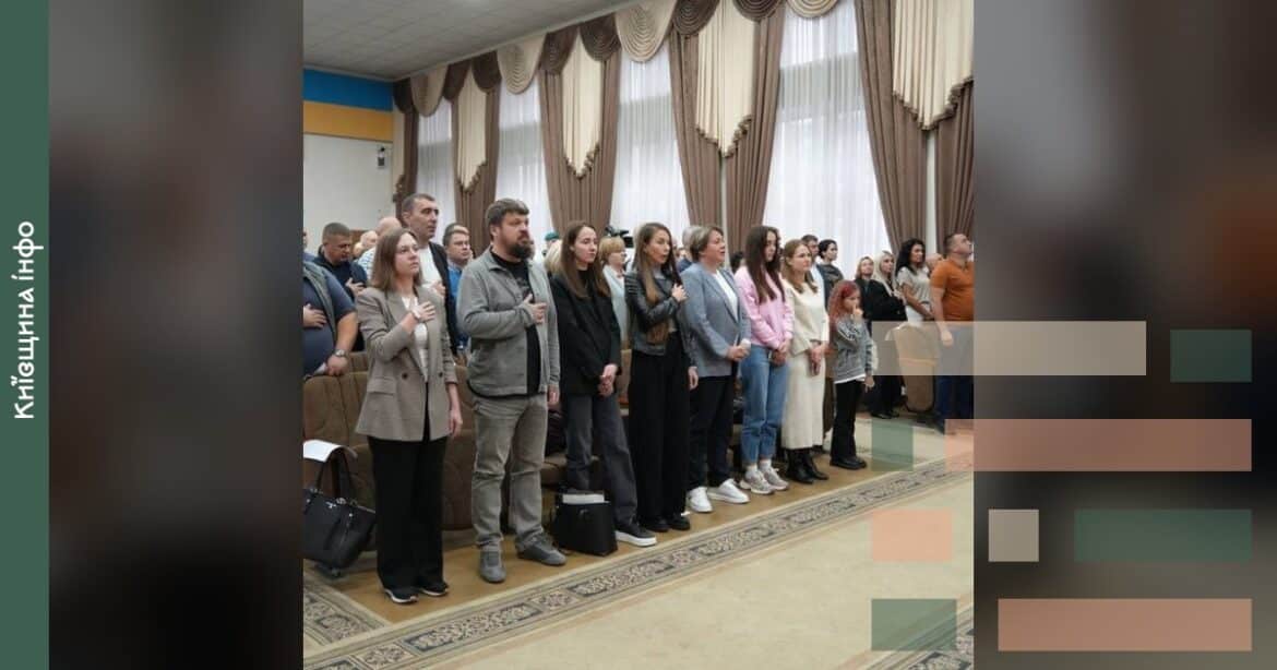 Учасники сесії Ірпінської міської ради стоять у залі засідань з декоративними завісами