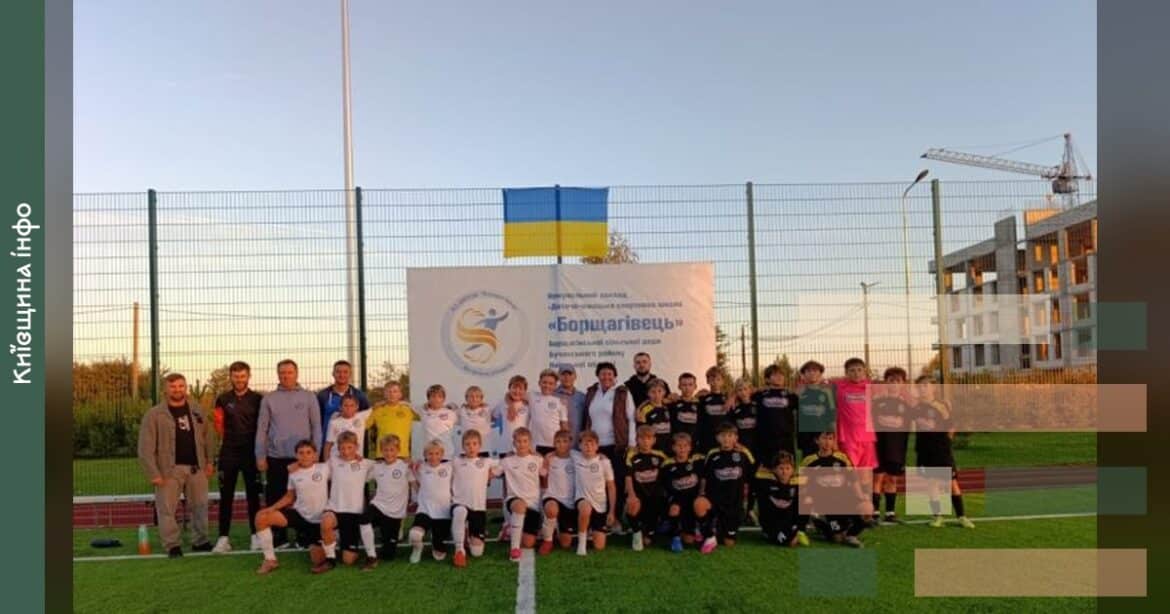 Юнаки та дорослі в спортивному форматі позують на футбольному полі з українським прапором і баннером ДЮСШ Борщагівець