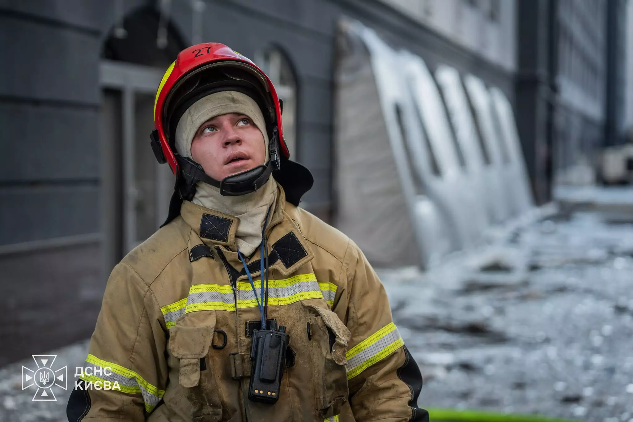 Молодий рятувальник у шоломі та захисному костюмі стоїть на фоні пошкодженого будинку після атаки