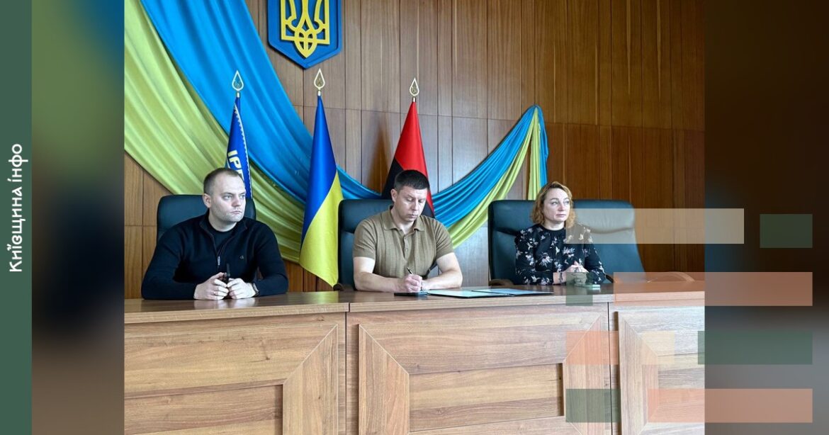 Три посадовці сидять за столом на засіданні в Ірпінській міській раді з українськими прапорами у фоні