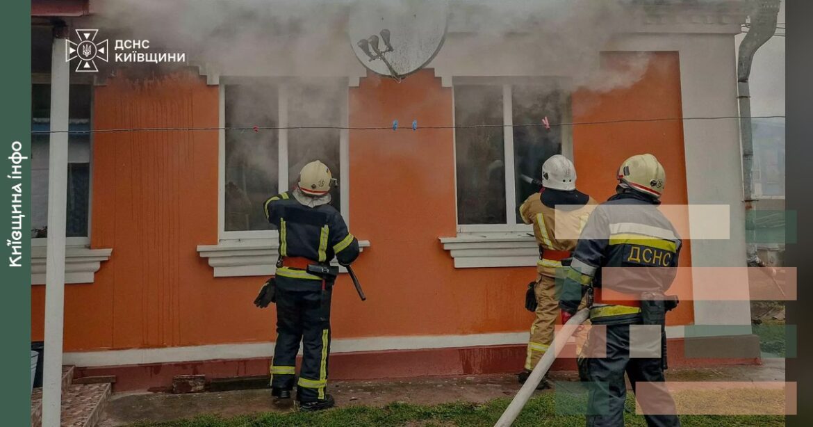 Рятувальники у касках і спецодязі гасять пожежу в оранжевому житловому будинку, з вікон йде дим