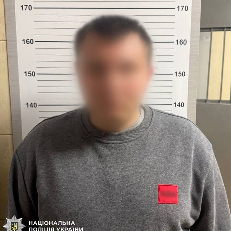 Чоловік у сірому светрі з червоною нашивкою стоїть на тлі ростоміра, обличчя на фото розмите, поліцейське фото з емблемою