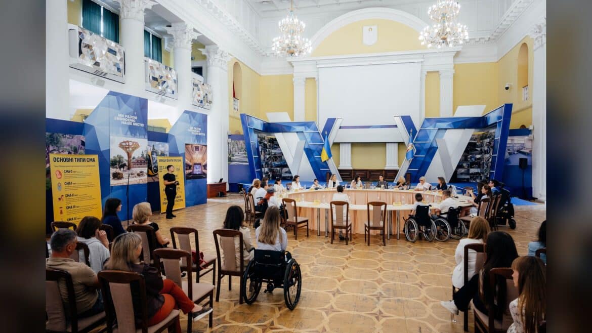 Конференція в залі з жовтими стінами, де люди, включаючи особу в інвалідному візку, сидять за столами під голубими