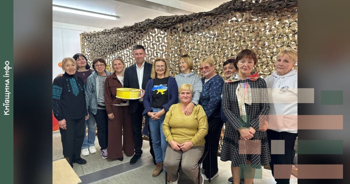 Група волонтерів осередку плетіння маскувальних сіток в Ірпені позує разом, чоловік в центрі тримає жовту тарілку на честь