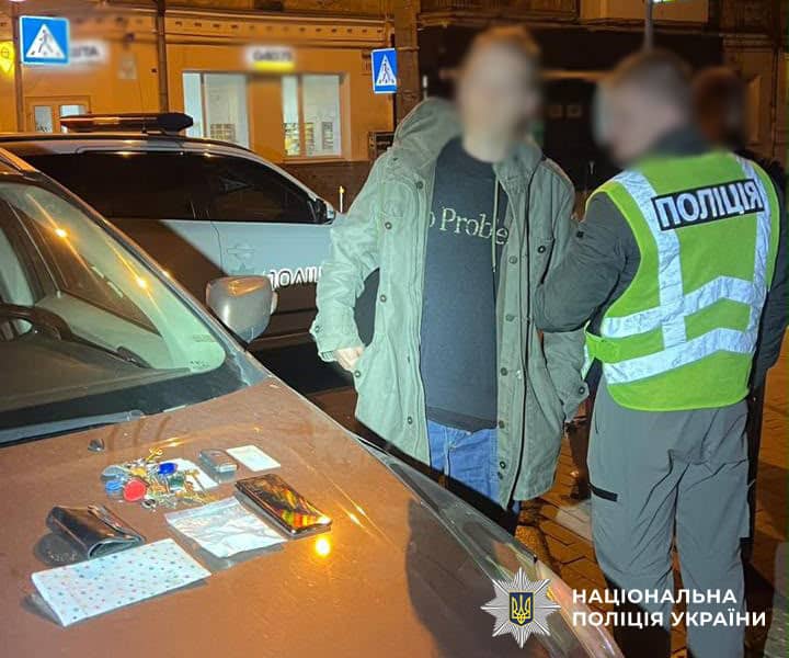Речі на капоті автомобіля, зокрема телефон, ключі та пакетики, на тлі поліцейського автомобіля вночі