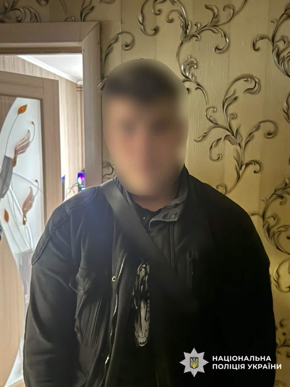 Молодий чоловік з розмитим обличчям у темній куртці стоїть на тлі шпалер із візерунком