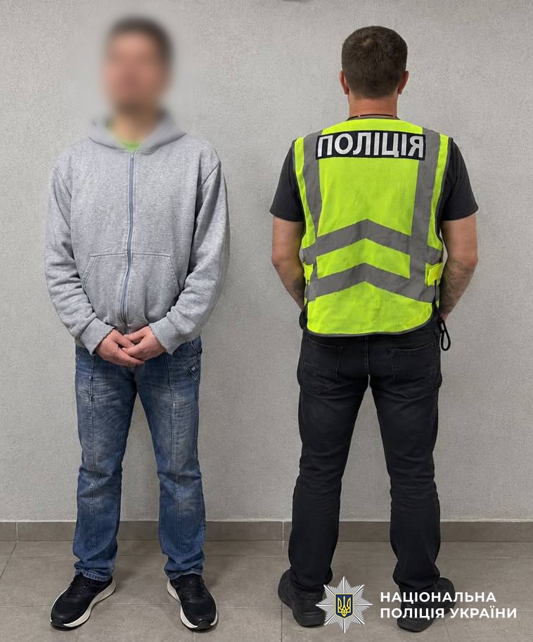 Чоловік у сірому худі з розмитим обличчям стоїть біля стіни поруч із поліцейським у жовтій жилетці