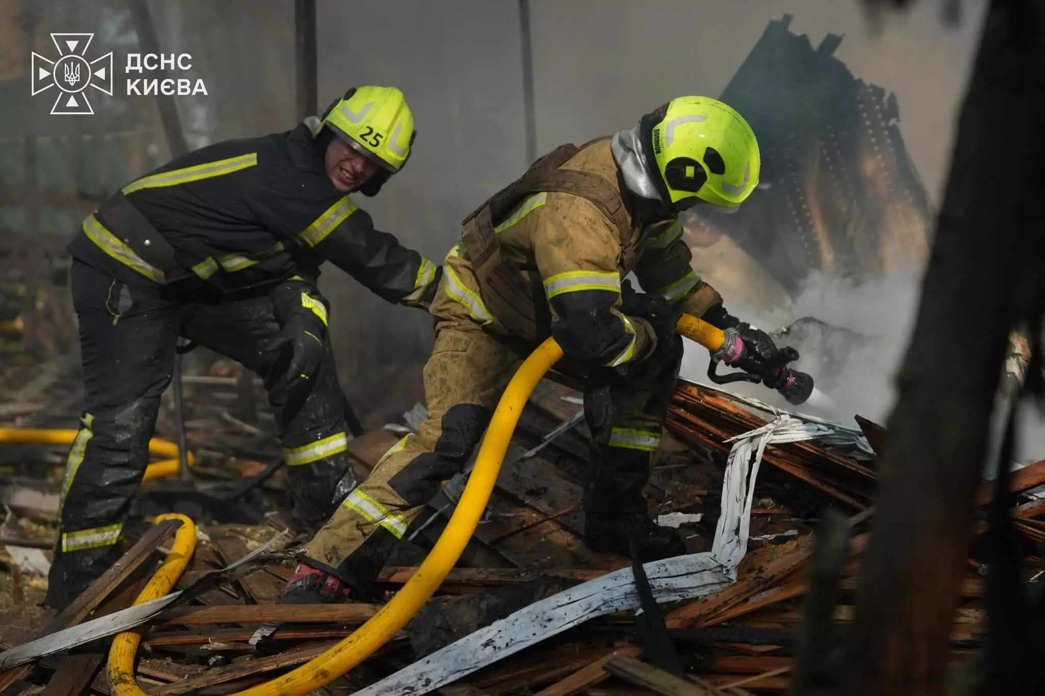 Двоє рятувальників у жовтих касках спрямовують пожежний шланг на горіючі деревʼяні уламки