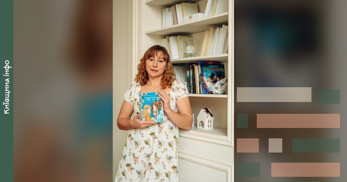 Письменниця у світлому плаття з квітковим принтом стоїть біля книжкової шафи з білими полицями