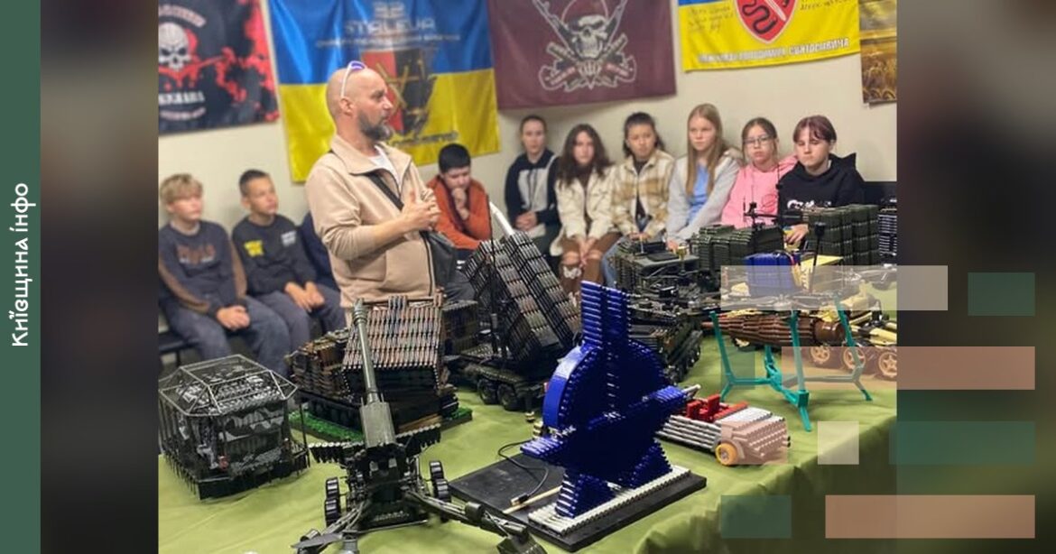 Чоловік презентує мініатюрні моделі військової техніки, виготовлені з гільз, групі учнів і молоді в залі з українськими