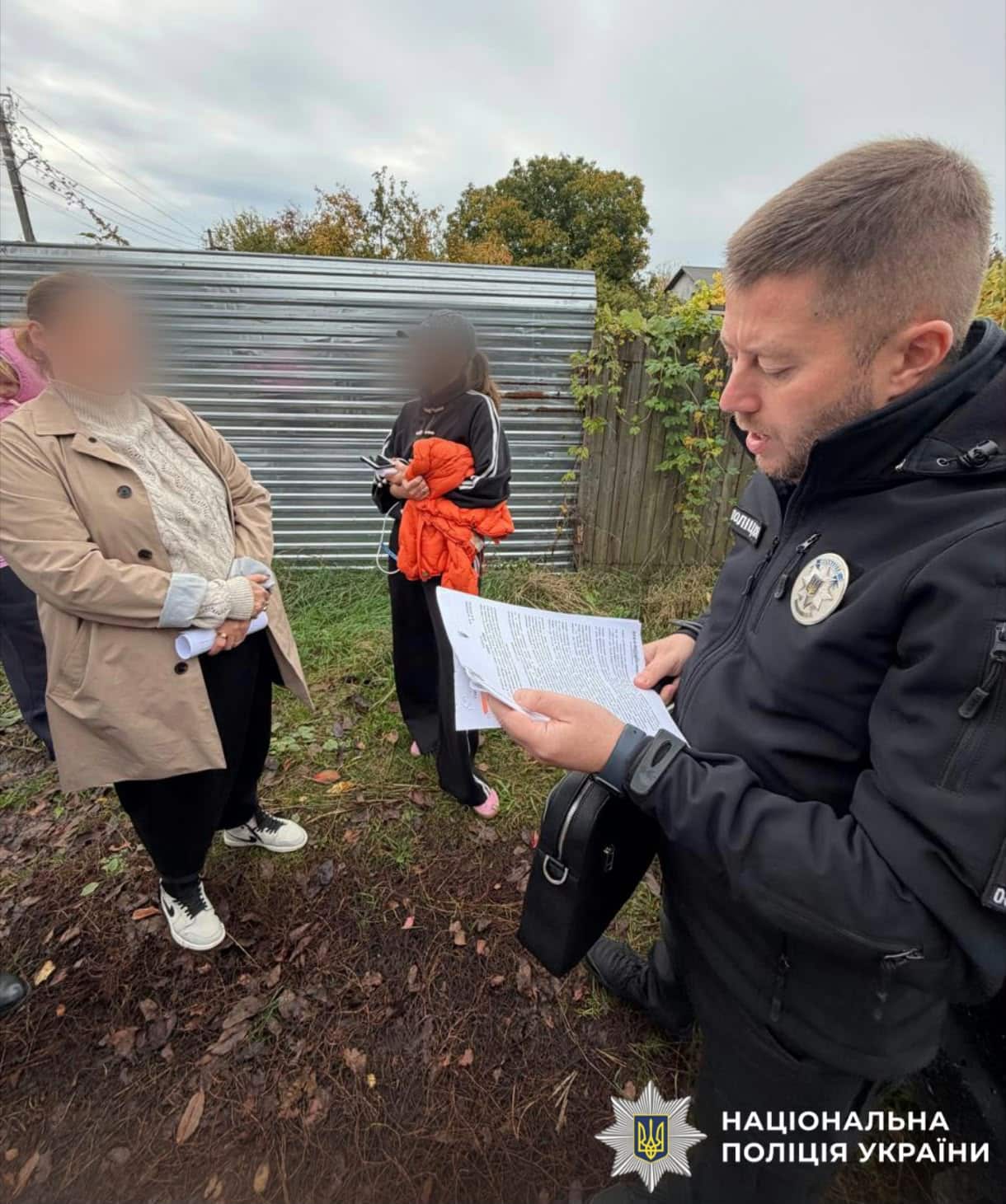 Поліцейський у чорній куртці читає документ на вулиці біля металевого паркану, поруч стоять дві жінки