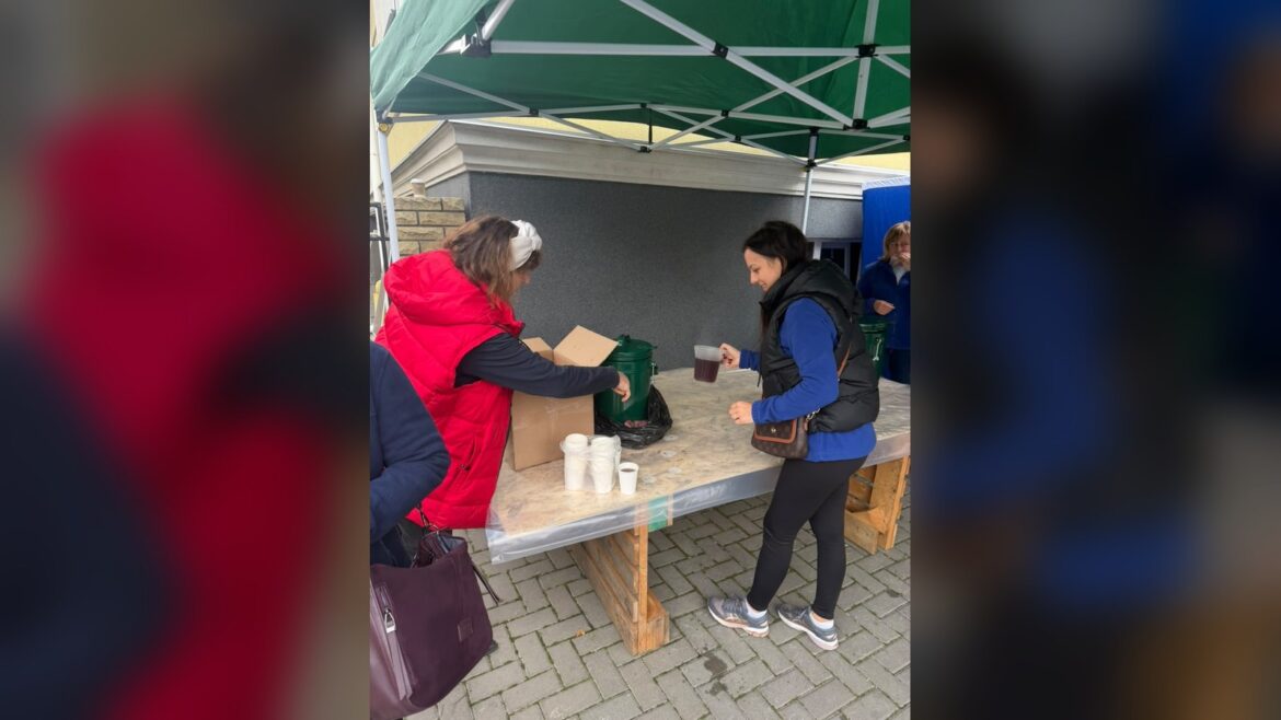 Волонтерка в червоній куртці видає гарячі напої з білих чашок постраждалій жінці на пункті допомоги під зеленим намітом
