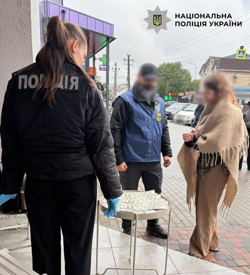 Поліцейська спиною до глядача, співробітник СБУ та жінка в бежевій шалі біля столика з грошима на вулиці