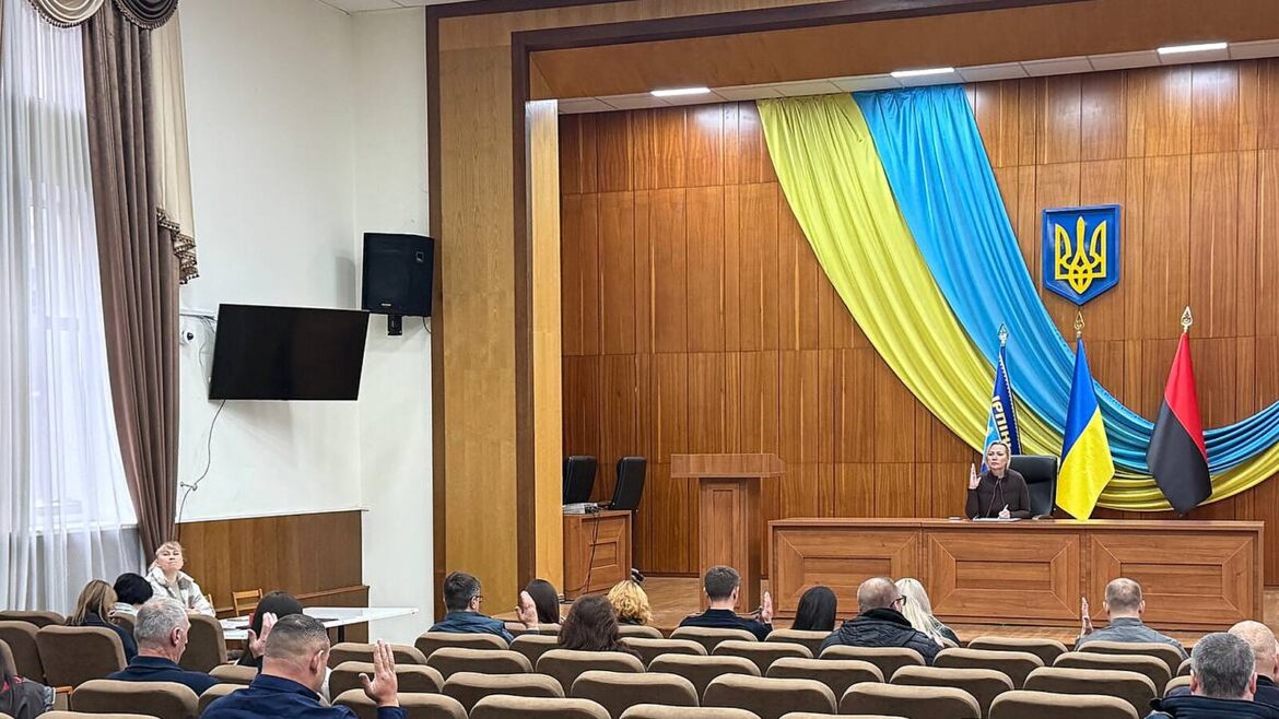 Засідання Ірпінської міської ради: кілька учасників піднімають руки на фоні синьо-жовтих прапорів і герба