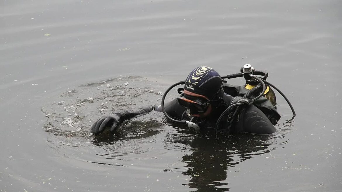 Водолаз у чорному гідрокостюмі з аквалангом на спині виринає з сірої води і тягнеться рукою вперед